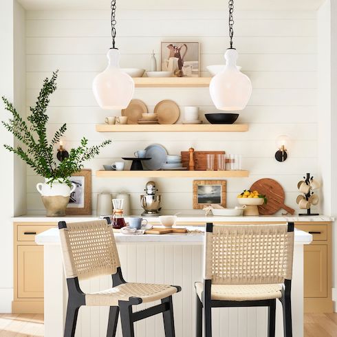 Two Flynn Milk Glass Pendants by Pottery Barn, featured in Home Network’s kitchen lights gallery hang in a small breakfast nook kitchen with wooden lower cabinets with black hardware, white marble countertop, three floating wooden shelves filled with bowls and plates, a small centre island with white panelling and two wicker and black bar stools