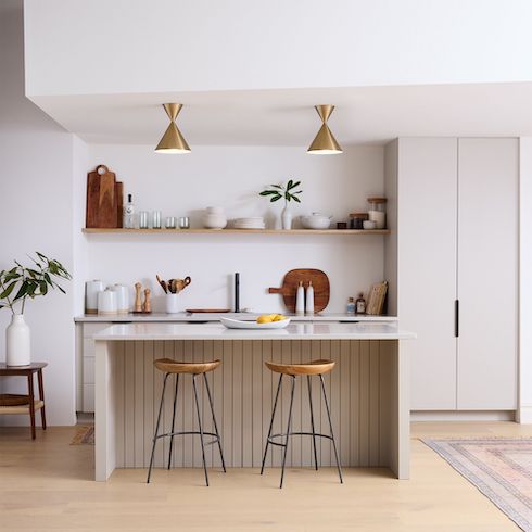 Two antique brass Warren Flushmount Ceiling Lights by West Elm featured in Home Network’s kitchen lights gallery shown in a small modern kitchen with white walls, a floating wooden shelf stacked with glasses, plates, bowls, glass storage and cutting boards, white stone countertops, centre island with beige wood panelling, a white fruit bowl and two wood and steel stools