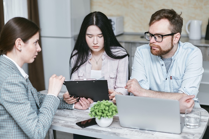 A woman in a business suit holding a tablet in front of a couple, negotiating an agreement.