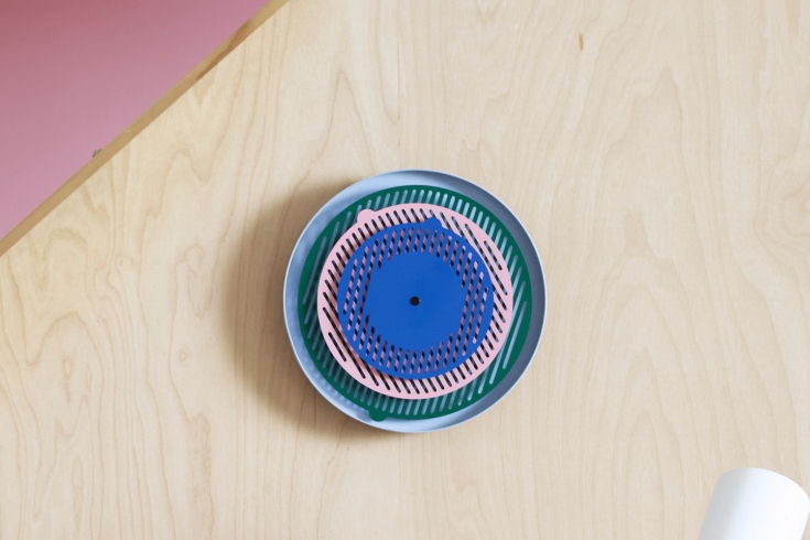 An abstract wall clock with three layers in dark green, light pink and dark blue.