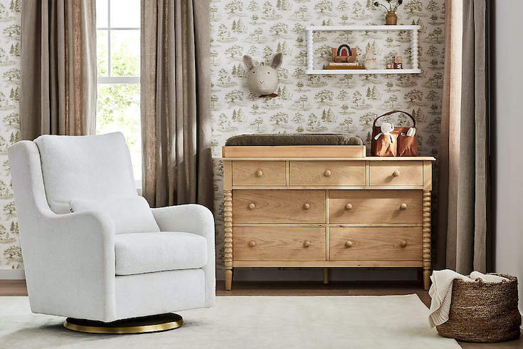 A Milo Faux Sherpa Nursery Swivel Glider Chair with Gold Base from Crate & Barrel shown sitting on a cream coloured rug in a nursery with green and cream pastoral wallpaper, café au lait linen drapes, a pine dresser change table with a brown changing pad and brown linen diaper caddy, a white display shelf and stuffed bunny head on the wall, and a jute basket with a white blanket sitting on the floor as part of Home Network.ca Nursery Trends for 2025