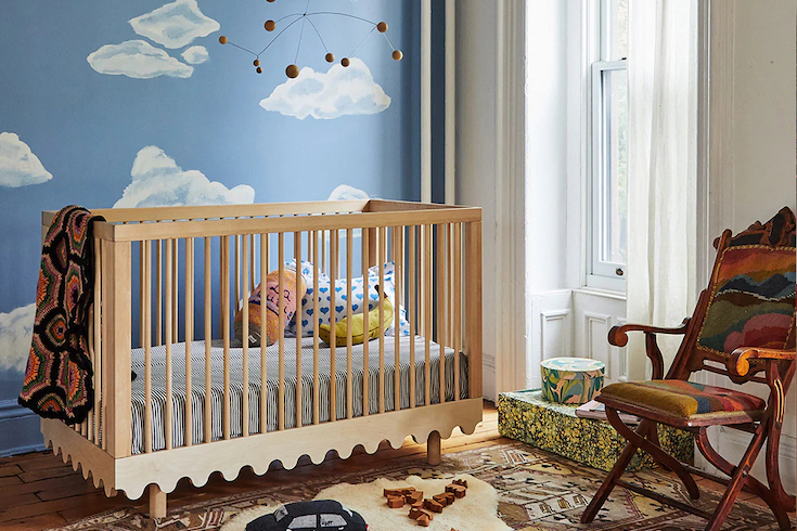 The Oeuf Moss Crib from Oeuf Canada, which features a bold-yet-simple wave motif and airy round spindles, sits in a charming modern nursery with a blue sky and white cloud wall mural, a modern metal and wood mobile overhead, a tall window with white gauzy drapes, two colourful boxes under the window, a brown and cream rug with a sheepskin rug on top, wooden floor, and an antique wood chair with colourful upholstery as part of Home Network.ca Nursery Trends for 2025