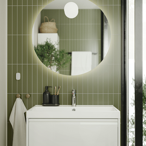 Sleek modern ÄNGSJÖN bathroom system from Ikea with a white vanity and sink, a large round backlit mirror, a black toothbrush set, two round towel hooks, and sage green subway tiles installed vertically shown in the Home Network gallery Unusual Bathroom Renovation Ideas That Will Be Popular In 2025