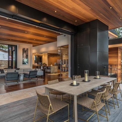 Home in Aspen, Colorado, with a large retractable wall surrounded by large clack tiles framing an interior with dark wood floors, living room with a white sectional couch and two grey armchairs, wood panelled ceiling with pot lights, a kitchen in the distance with a large marble topped island, and deck with Mbrico Tiles and a large dining table with woven dining chairs