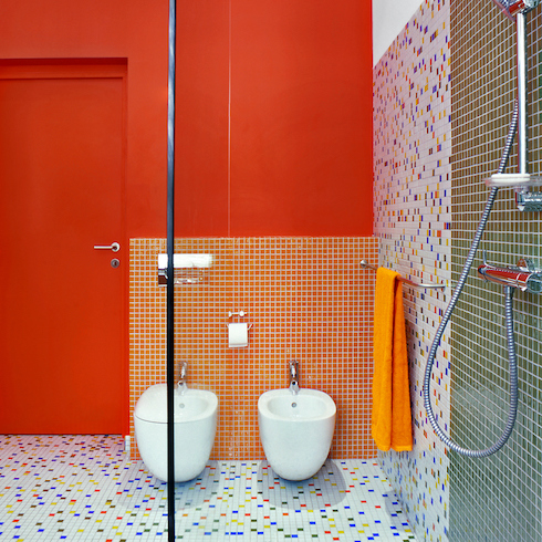 Colourful bathroom with white and multicoloured tile floor, red wall, white bidet and toilet, and a metal towel rack with an orange towel shown in the Home Network gallery Unusual Bathroom Renovation Ideas That Will Be Popular In 2025