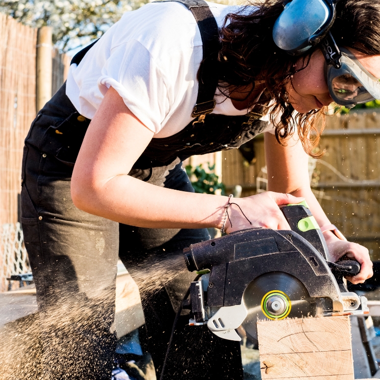 Woman wearing protective goggles and ear protectors holding circular saw, cutting piece of wood on building side.