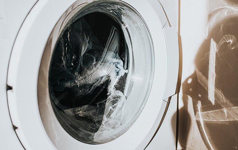 Close-up on a white washing machine with a glass front, as clothing spins around mid-cycle. The glass casts a shadow on the wall