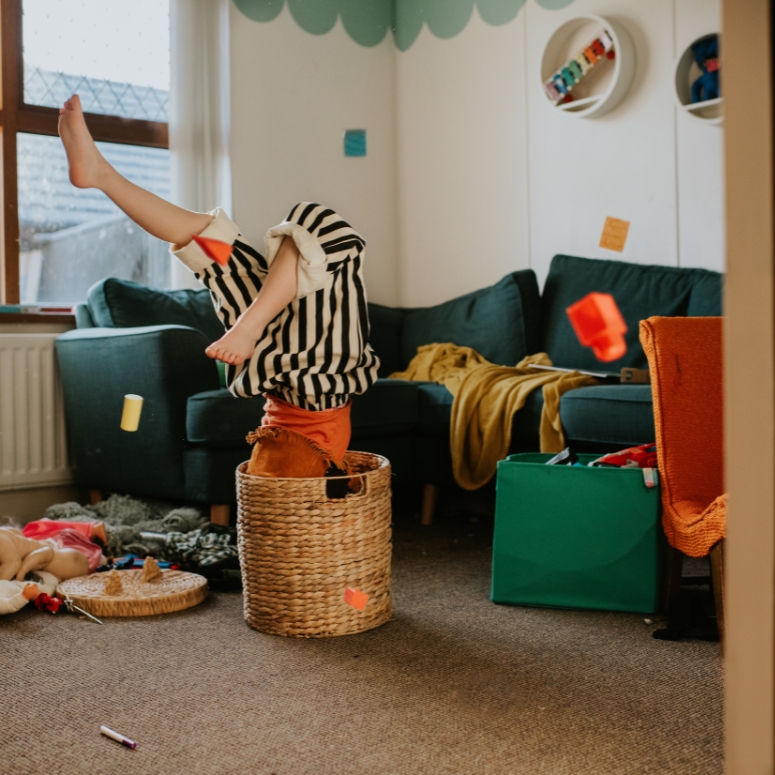 A little girl mischievously climbs inside a small wicket basket.