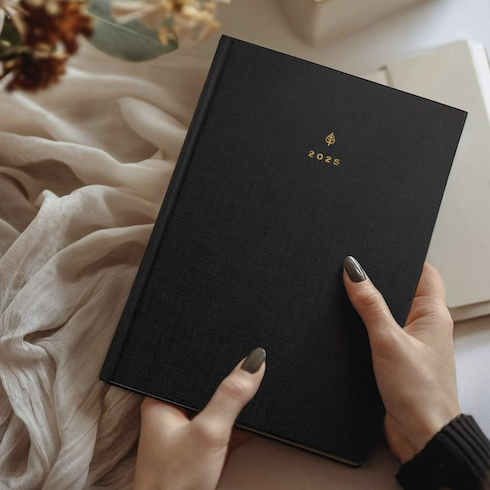 Manicured hands hold a new black 2025 Hemlock & Oak planner above a desk decorated with holiday decor as part of HGTV's 10 Gift Ideas for the Women in Your Life