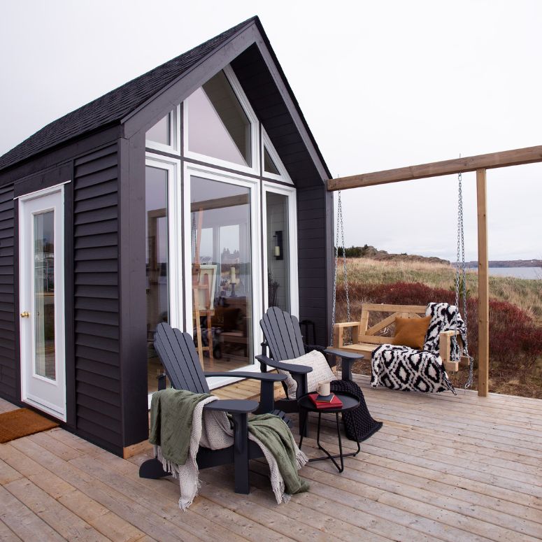 Dark gray studio by the water with a deck, dark muskoka chairs and a wooden bench swing