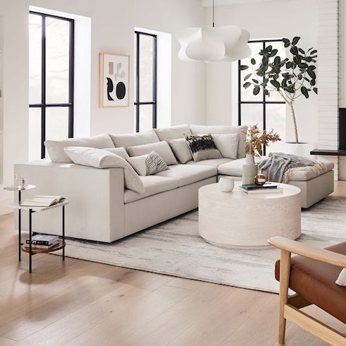 Cloud couch dupes featuring a Harmony Modular 3-Piece Ottoman Sectional in a big bright white living room with big black framed windows, light wood floors, modern art on the walls, a large white and grey rug, round coffee table, and a large potted plant in a white pot