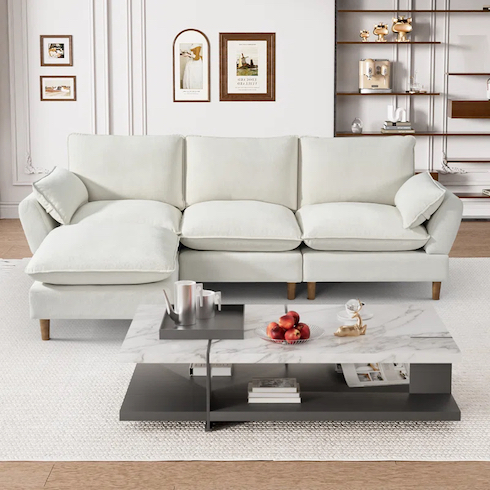 Cloud couch dupes featuring a Tetiana L-shape Upholstered Cloud Modular Sofa in Beige Chenille from Wayfair in a white living room with a large white area rug, a modern marble and black coffee table, and white wall with moulding and hung art