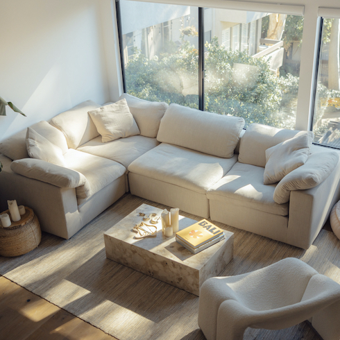 Cloud couch dupes featuring Eternity Modern's EM Sky Sofa modules put together in a living with a wall of windows looking out to a garden below, wood floors, a beige area rug, a curved occasional chair, and stone coffee table piled with books and candles