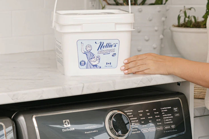 Nellie's Laundry Soda Concentrated Laundry Detergent as featured in Laundry Mistakes on HGTV Canada sits on top of a washing machine countertop with a hand holding the bucket