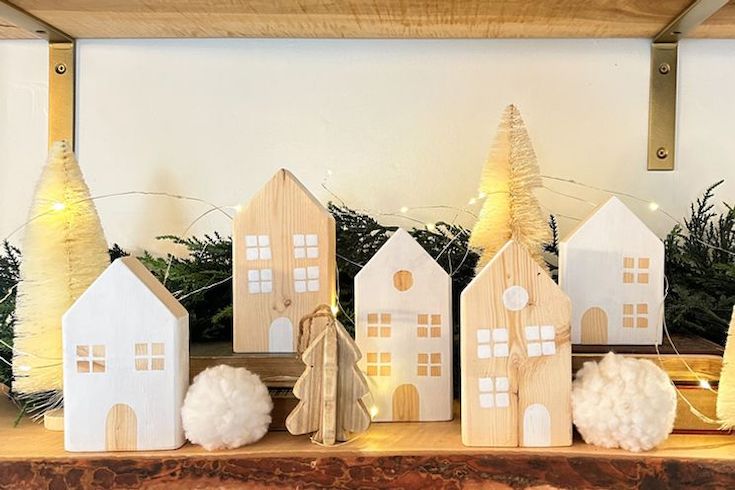 More Sustainable Holidays featuring a wooden DIY Christmas Village by Courtney Ryall sitting on a shelf lit by white fairy lights