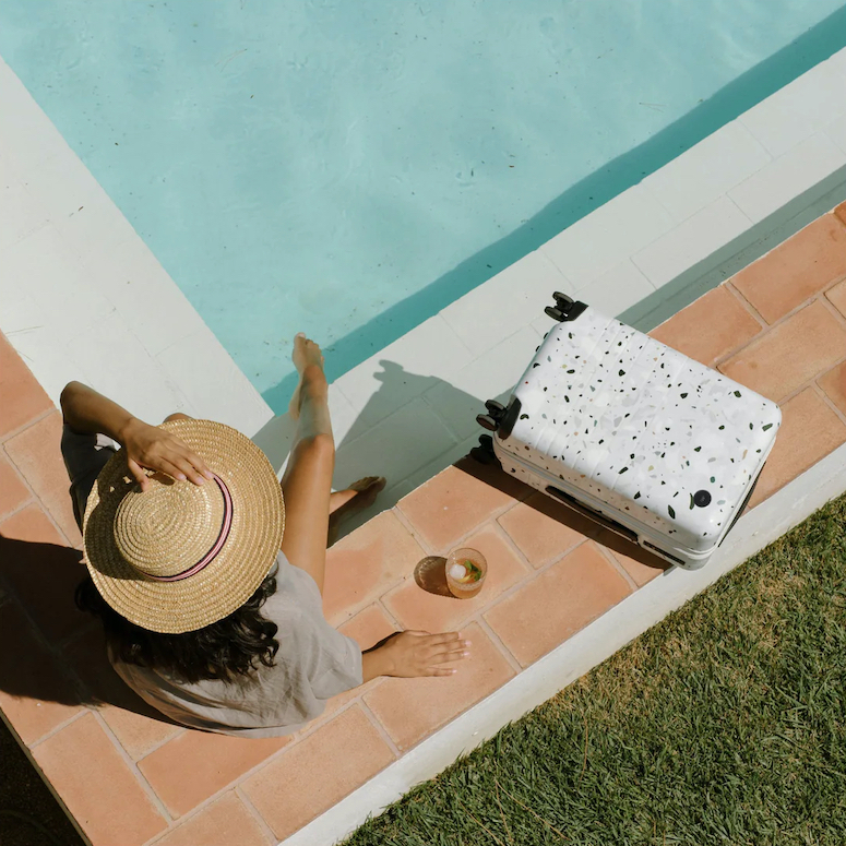 Monos Carry-On Plus Limited Edition Terrazzo aerial view beside a pool with a woman sitting there in a straw hat