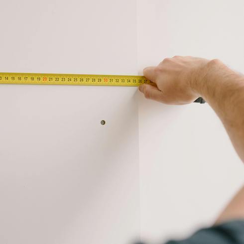 Person holding up a yellow measuring tape to a white wall measuring to make sure there is enough space before storing holiday decorations for Home Network