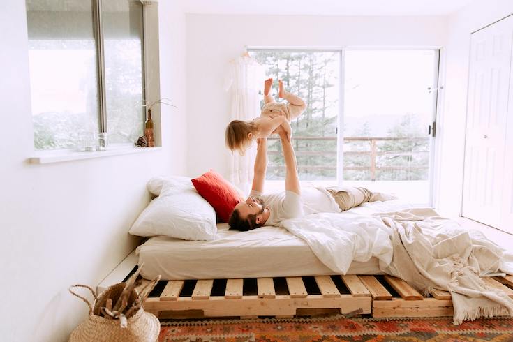 Man and child play on a platform bed in a bright white bedroom for Home Network When You Should Replace Your Mattress