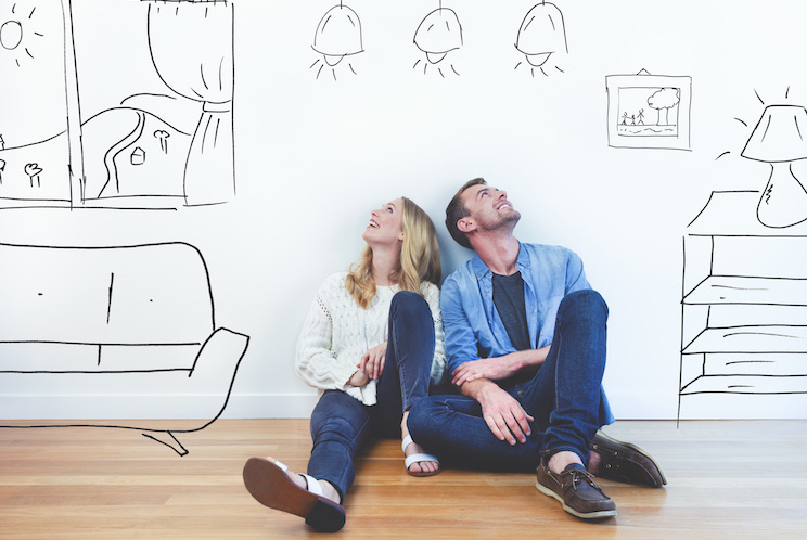 Couple dreaming of their new house. They are sitting on a wooden floor imagining their new home with furniture in it. The house is currently empty. They are booth looking up and smiling happily.
