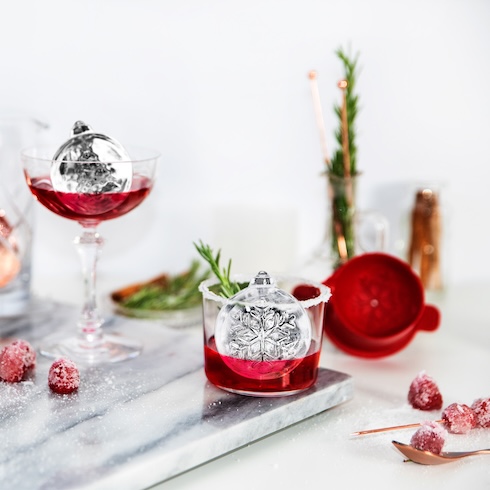 Two glasses are filled with a red drink and ornament-shaped ice cubes on a festive table.