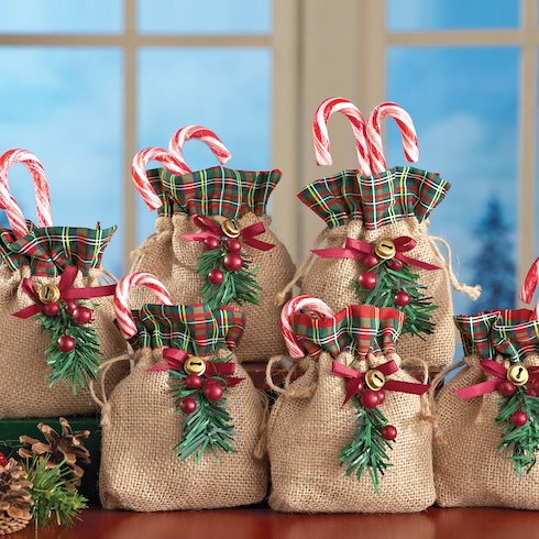 Several small burlap-and-plaid gift bags are arranged in front of a window. Each bag has a bell at the front and has red-and-white candy canes sticking out from the top.