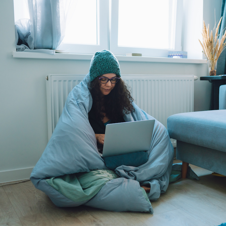 Middle age business woman work at home sitting on a floor next to heater wrapped in blanket and wearing woollen hat