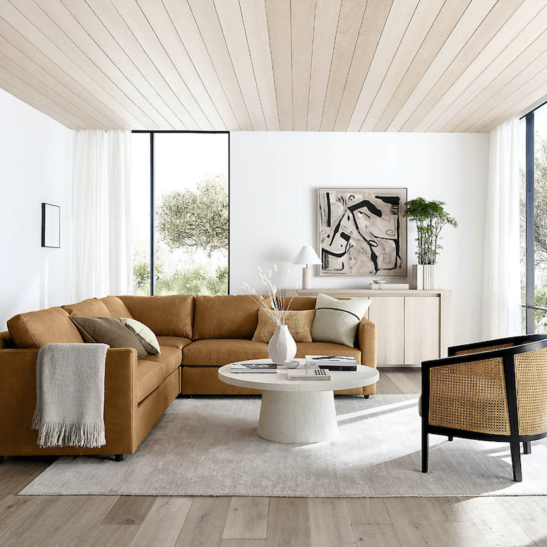 Crate & Barrel Willy White Concrete Pedestal Coffee Table designed by by Leanne Ford in a bright modern living room with a brown sectional couch, white rug, white oak floors, white walls, batten ceilings, and a black cane chair