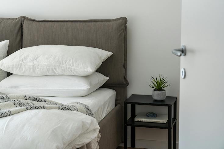 Beautiful modern bedroom with a grey fabric headboard, white linens and pillows and a small bedside table for 10 Hidden Spots That Need a Deep Clean in Your Bedroom by HOME Network