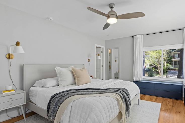 A simple bedroom with bedside table, bed with white, grey and yellow linens, white walls, a ceiling fan, wooden floors and windows