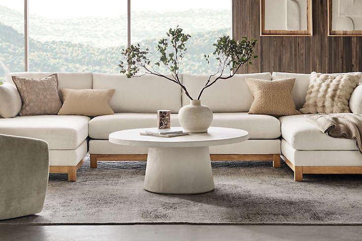 Crate & Barrel Willy White Concrete Pedestal Table by by Leanne Ford sits in a modern living room with a vase, books and candle, sitting on top of a large area rug with a huge white sectional couch and large windows looking out over a mountain view