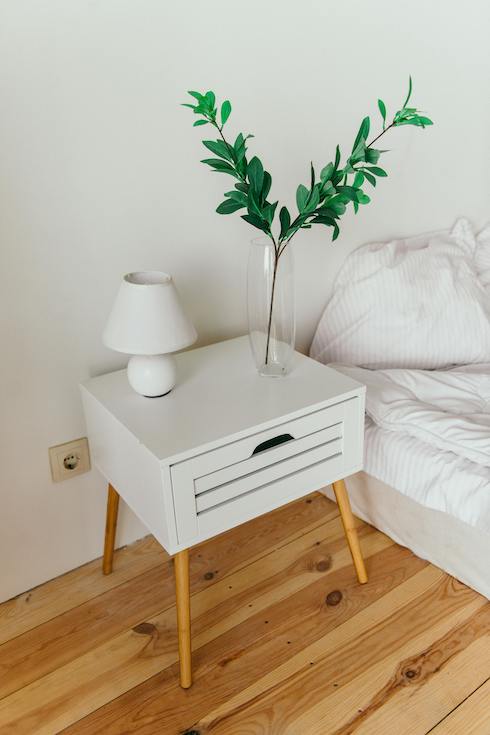 Mid century modern bedside table in white lacquer and wood sits on a wood floor in a simple bedroom with a white bed for Home Network's 10 Hidden Spots That Need a Deep Clean in Your Bedroom