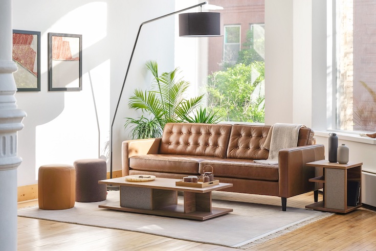 EQ3 Plank Rectangular Coffee Table in a masculine living room with a cream coloured area rug, a tufted leather couch, two leather ottomans, a large gooseneck floor lamp, large potted plants and huge floor to ceiling windows showing how to section a coffee table in a post of Home Network on styling coffee tables