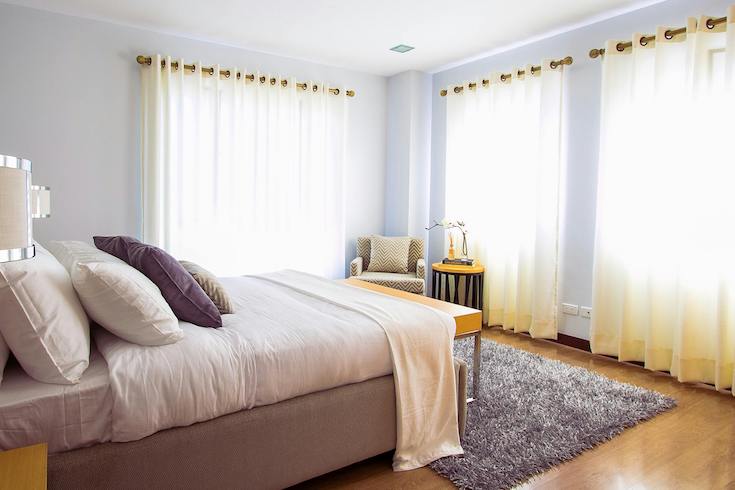 Lovely open, airy bedroom with wood floors, a bed with pink and lilac linens, blue grey walls, and light yellow curtains drawn over large light filled windows