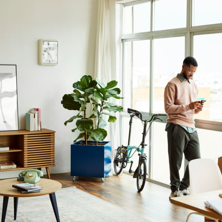 A man on his phone while leaning on window of his spacious home office featuring plant, a bike and wooden coffee table and chest.