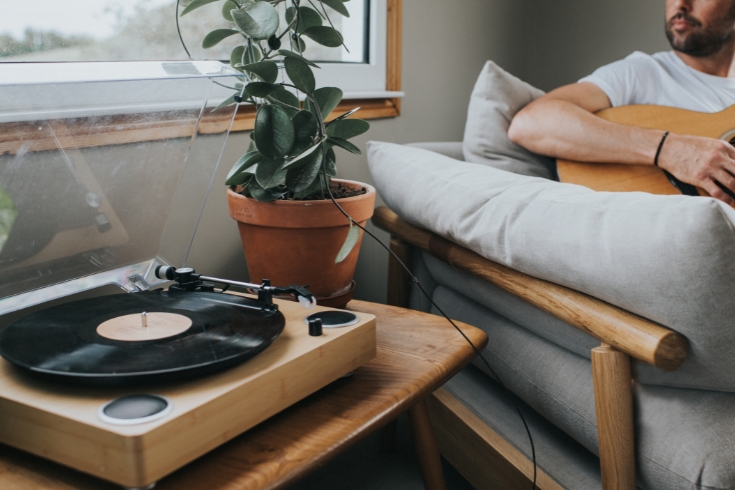Man sits on a comfortable sofa in a stylish living room playing a guitar. Focus is on the modern record player that sits on the table next to him. 