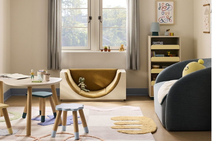 Eva Chen Arched Reading Nook with Bookcase for West Elm sits on the floor of a kids room with a table and chairs, and a small couch in a sun filled room with window.