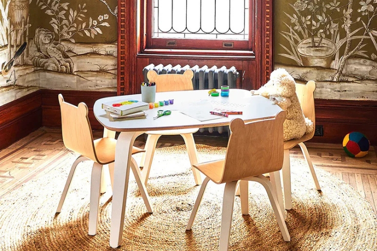 Oeuf Canada Play Table with rounded edges sits on a woven rug in a sun filled kids bedroom and playroom with dark wood trim and patterned green wallpaper and is set up with art supplies and surrounded by four bear chairs