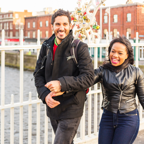 A smiling man and a woman walk arm in arm across the Ha'penny Bridge in Dublin, Ireland.