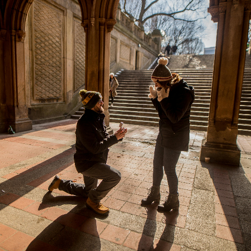 A man on one knee proposing marriage to a woman at Bethesda Terrace in Central Park, New York City