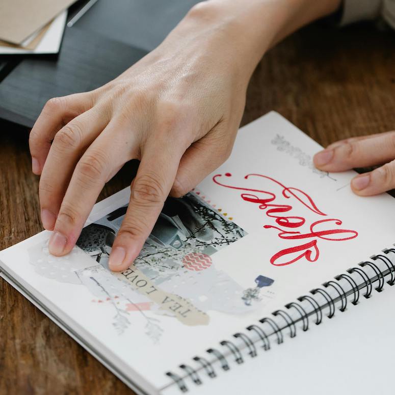Woman Making a Scrap Book and gluing down pictures to a page of a junk journal onto top of a wooden desk beside a black laptop as part of the post How to Start a Junk Journal on Home Network
