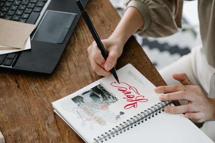 Woman Making a Scrap Book and writing in the pages of a junk journal onto top of a wooden desk beside a black laptop
