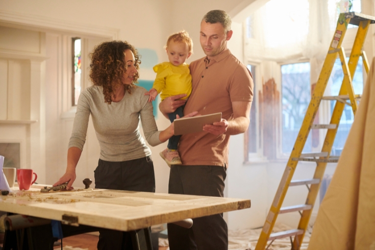 A couple, with the man holding a toddler, looking at documents in a room under renovation.