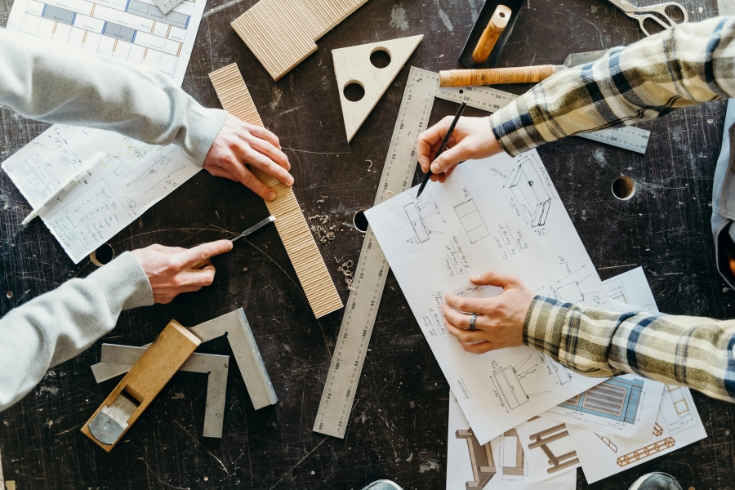 Hands of two people sketching blueprint and writing down measurements with multiple rulers laid out on table.