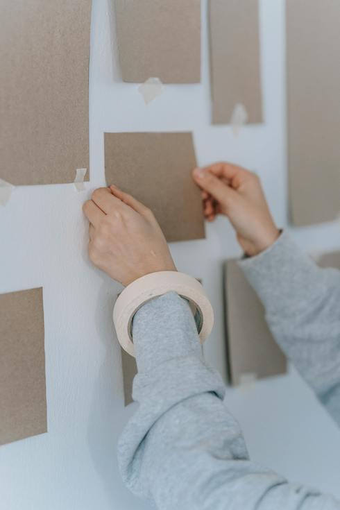 Person in Gray Long Sleeve Shirt Holding A Cardboard frame template up to a Wall to make a gallery wall