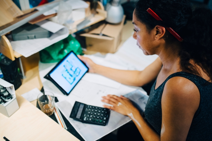 Woman analyzing blueprint on screen while doing calculations on paper with a calculator.