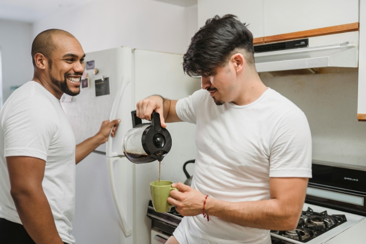 Two men smiling in the kitchen, one opening the fridge while the other pouring coffee