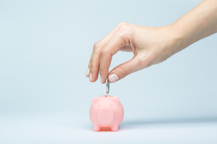 A hand drops a coin into a tiny pink piggy bank.