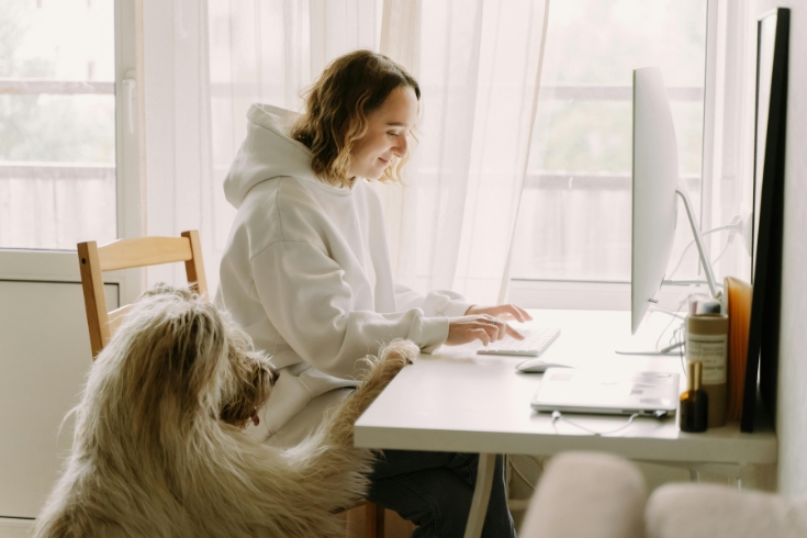A young woman types on computer while a dog sits beside her.