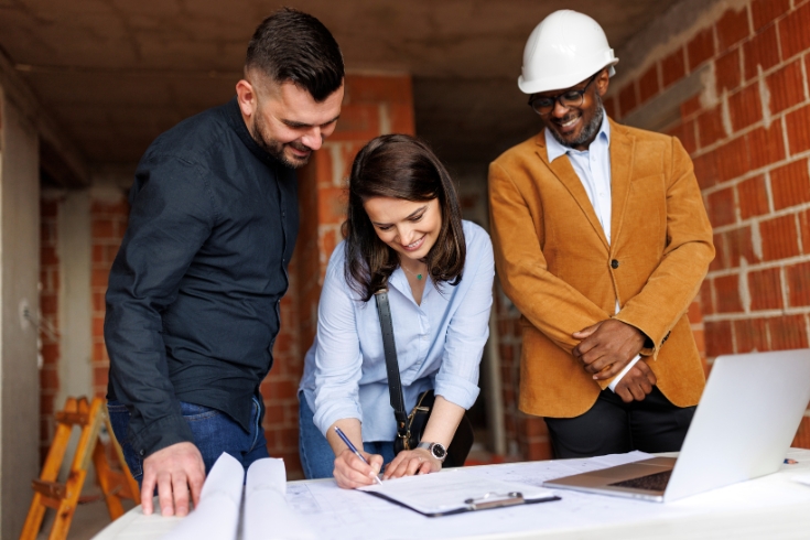 A man, a woman and a man wearing a construction helmet stand smiling, while the woman signs on a document.