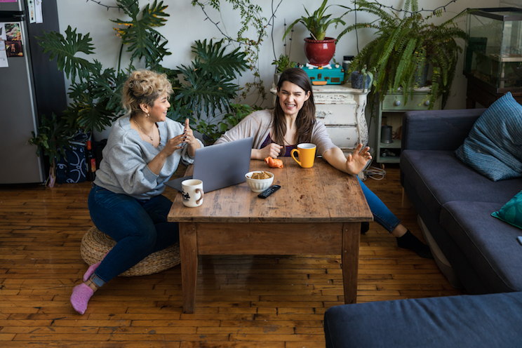 Happy friends talking while sitting with coffee mugs and laptop at table